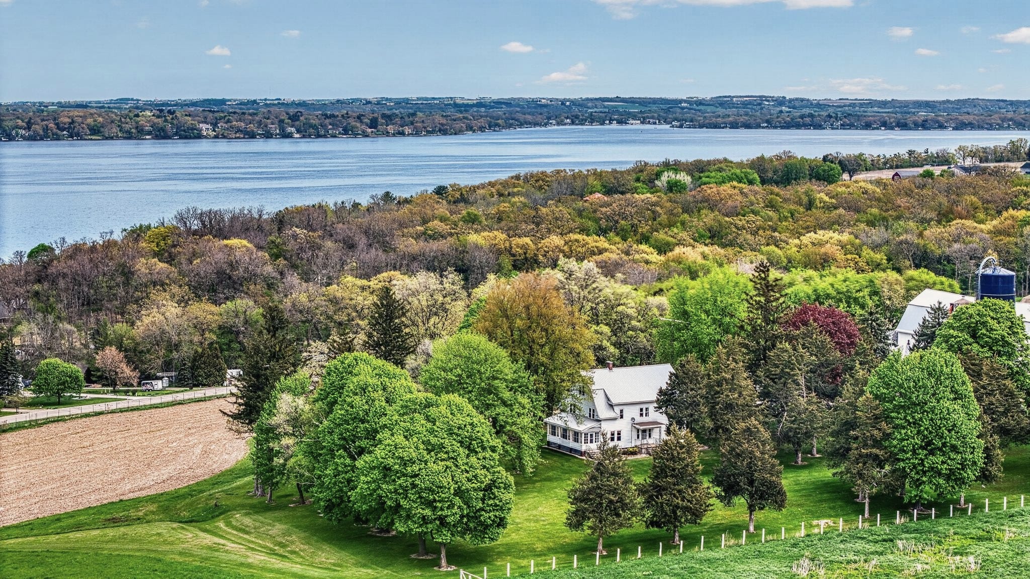 Drone view of Hilltop Haven, a white farmhouse among trees and fields, with Green Lake beyond.