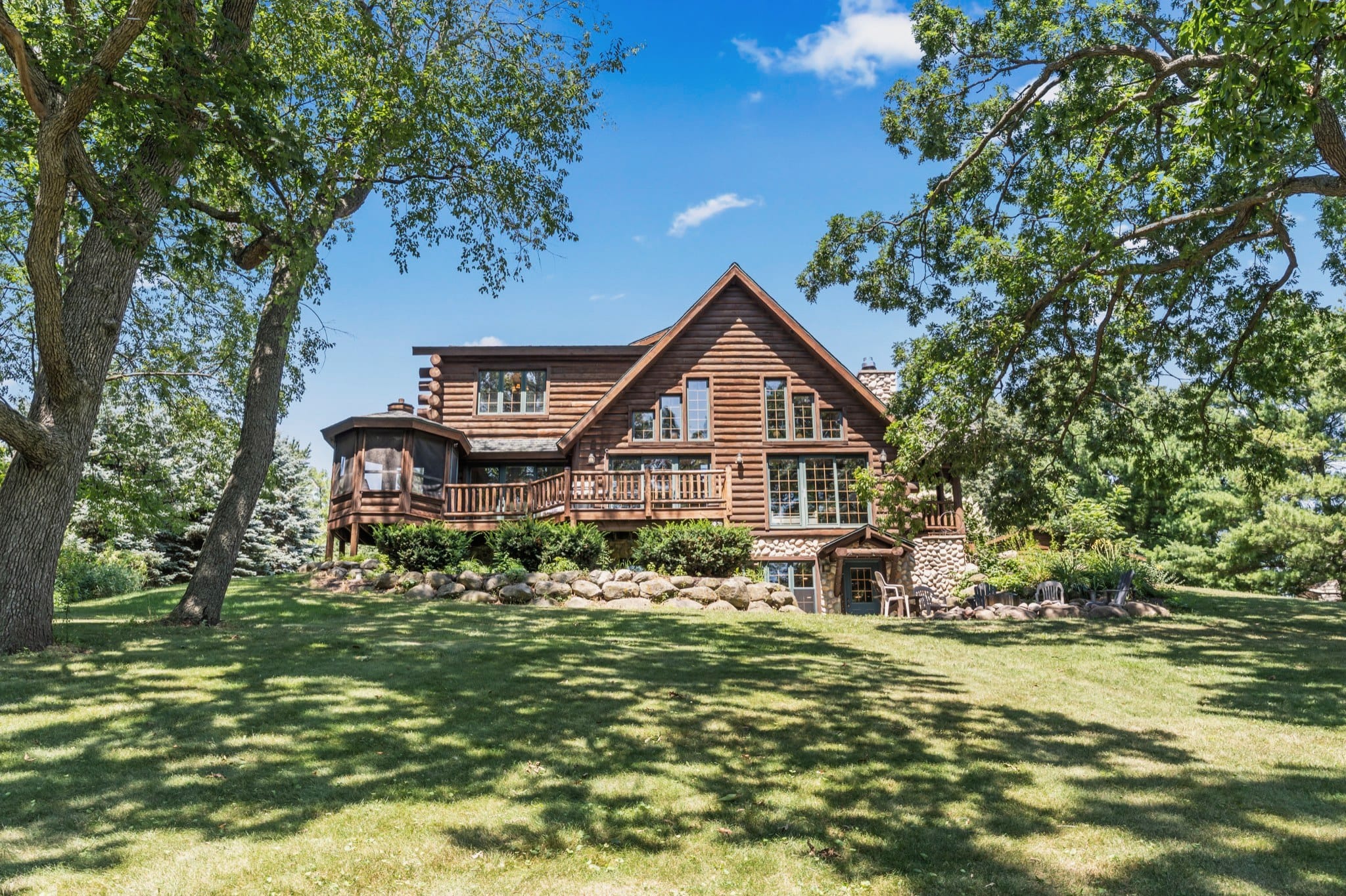 The back of the log cabin seen across a shaded lawn between two tall trees: a gable of tall windows, a long deck with a log railing and a screened gazebo at its end, and a fieldstone lower level with a walkout door and patio chairs.