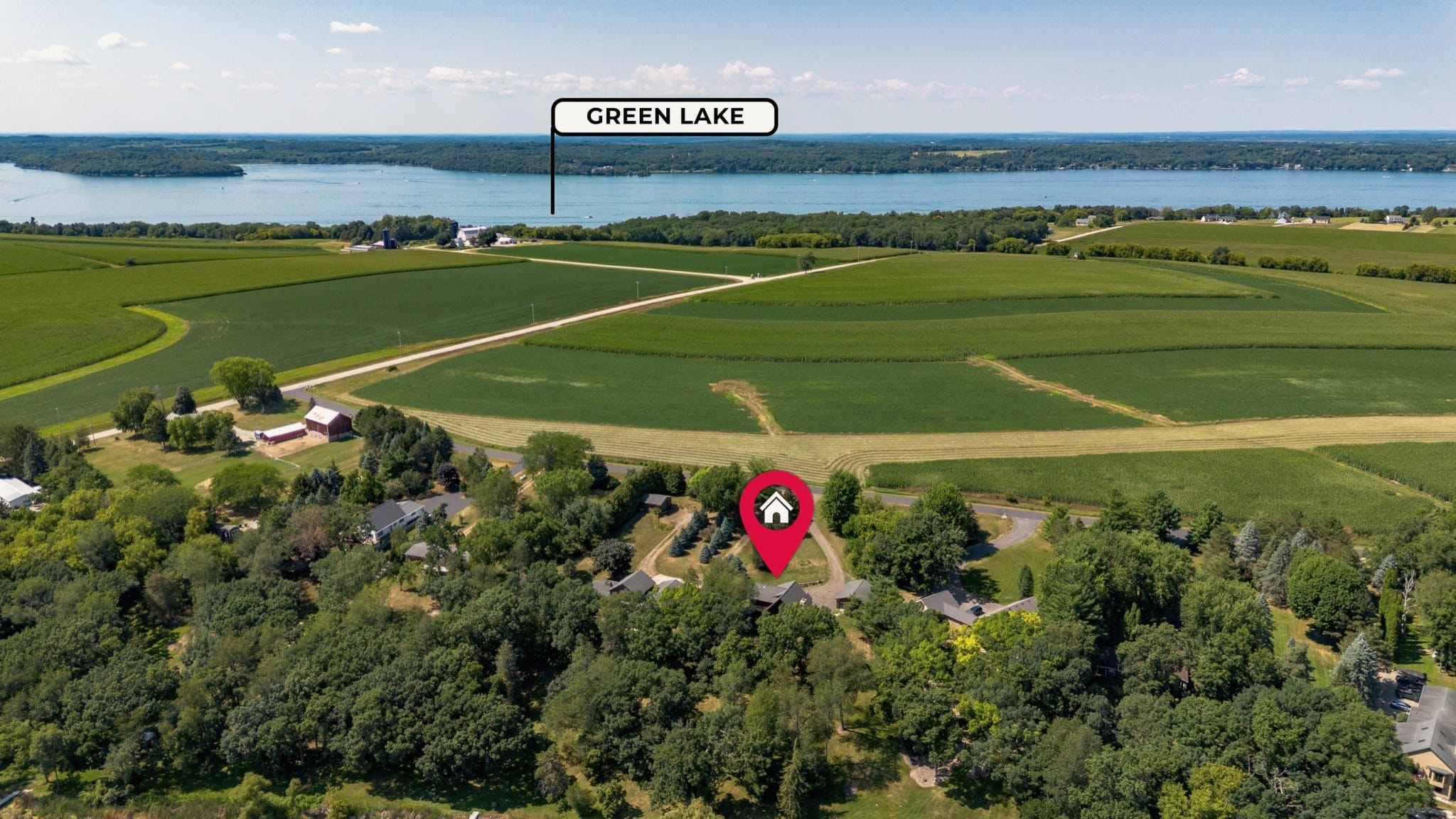Drone view of the trees around the cabin, marked by a red map pin added to the photograph, looking across farm fields to Green Lake on the horizon, named by a label on the photograph.