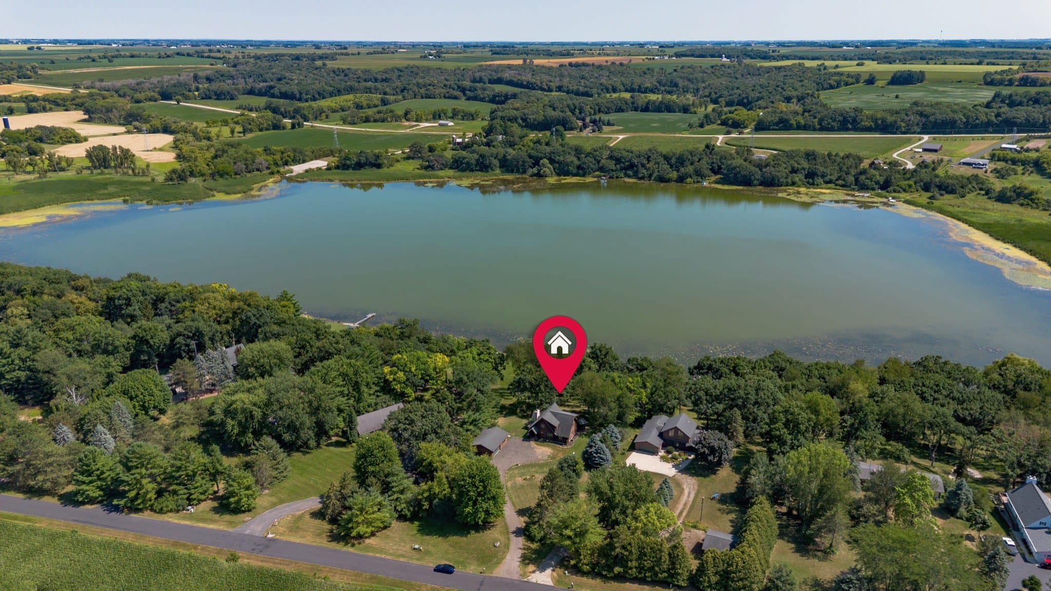 Drone view of the cabin among trees on the shore of Big Twin Lake, marked by a red map pin added to the photograph, with woods and farm fields beyond the lake.
