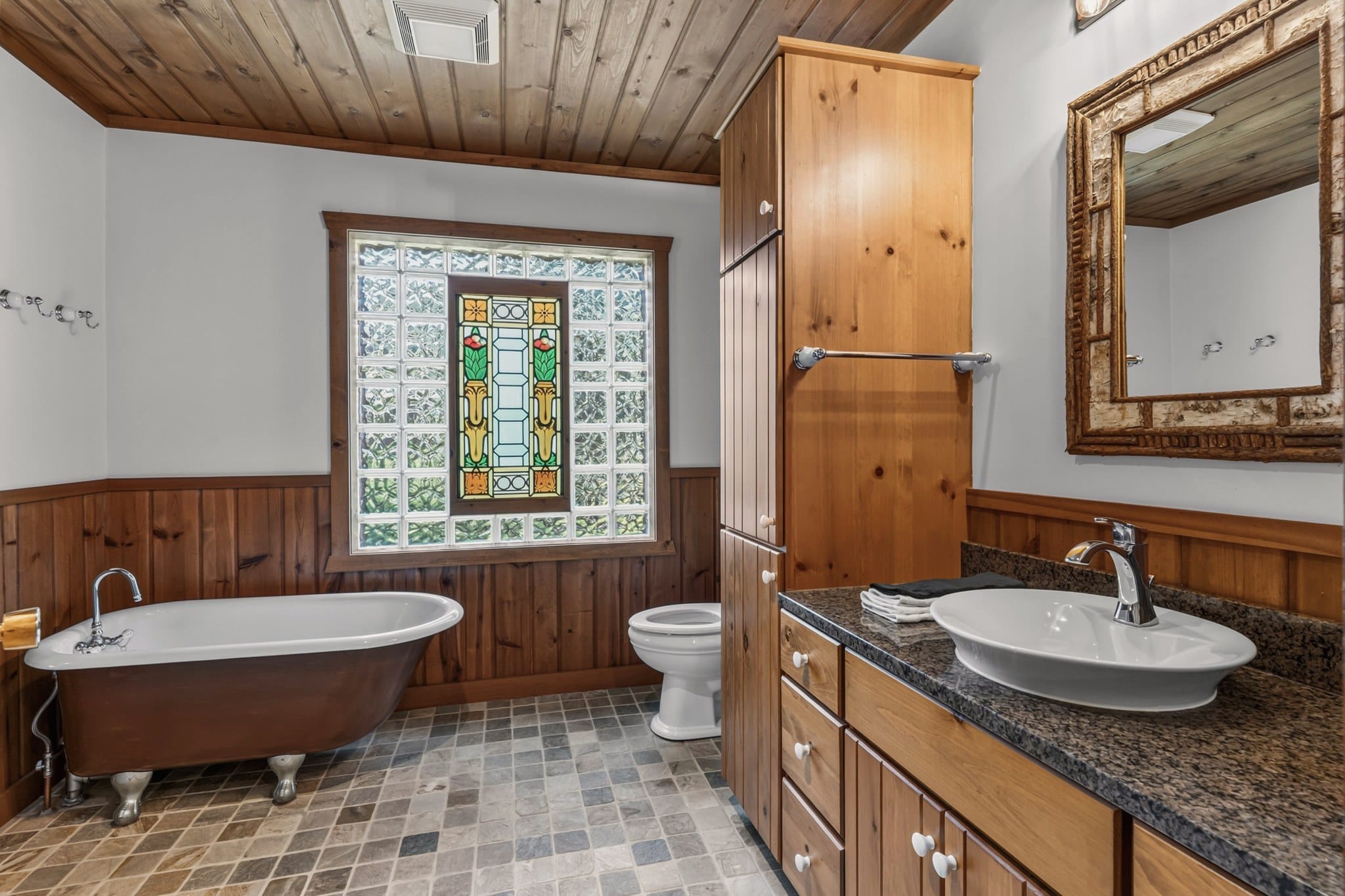 A bathroom with a plank ceiling and a tiled floor: a clawfoot tub under a stained glass window set in glass block, a toilet, a tall pine cabinet, and a white vessel sink on a dark granite counter below a mirror in a rustic wooden frame.