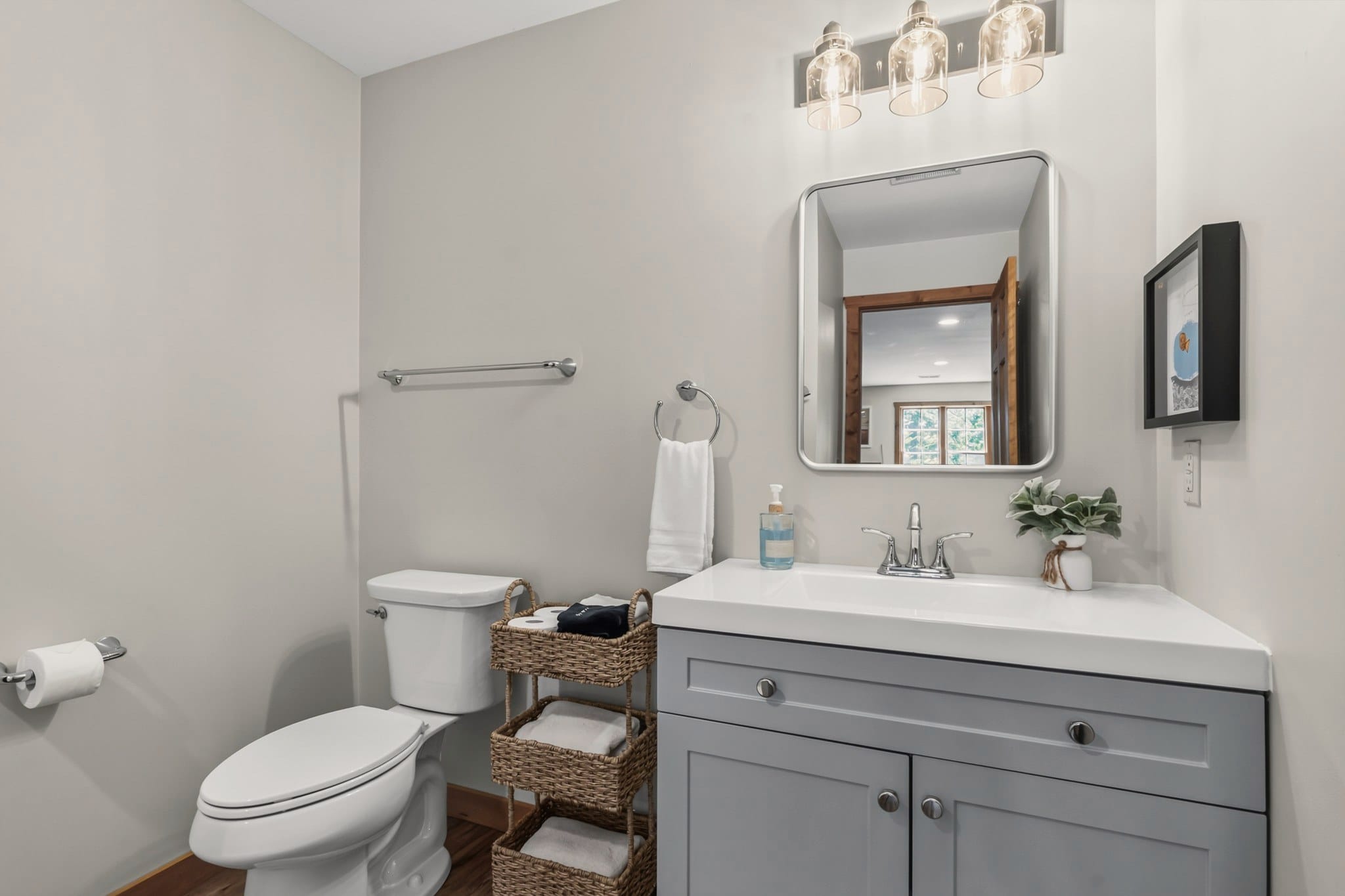 A bathroom with pale grey walls: a toilet, a woven basket stand of rolled towels, and a grey vanity with a white sink under a rounded mirror and a three-light fixture.
