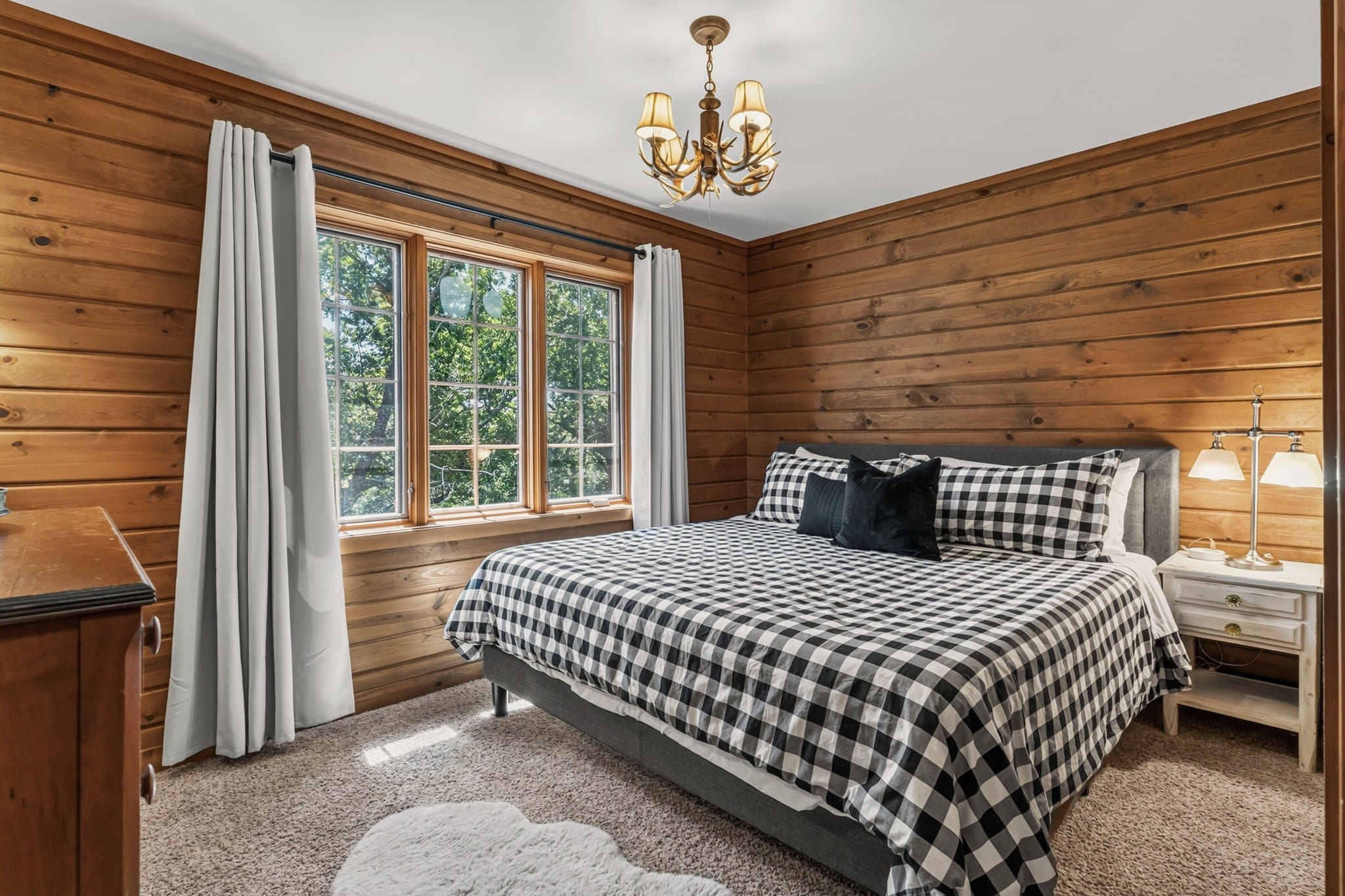 A bedroom with pine plank walls and a carpeted floor: a king bed in black and white check with an upholstered grey headboard, an antler chandelier, three windows into the trees with grey curtains, a white nightstand and a dresser.