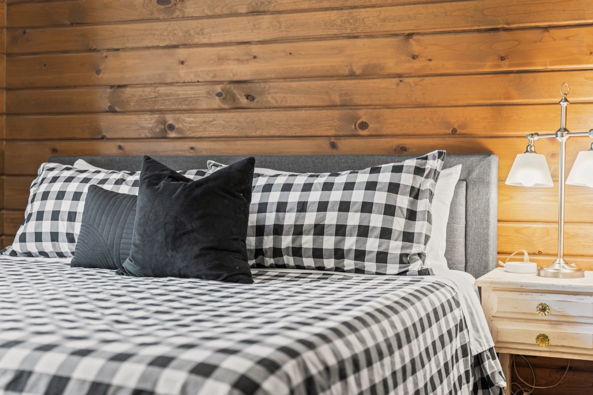 Close view of the check bedroom's bed: black and white check pillows and cover, two black cushions and a grey upholstered headboard against the pine wall, beside a white nightstand with a two-arm lamp.