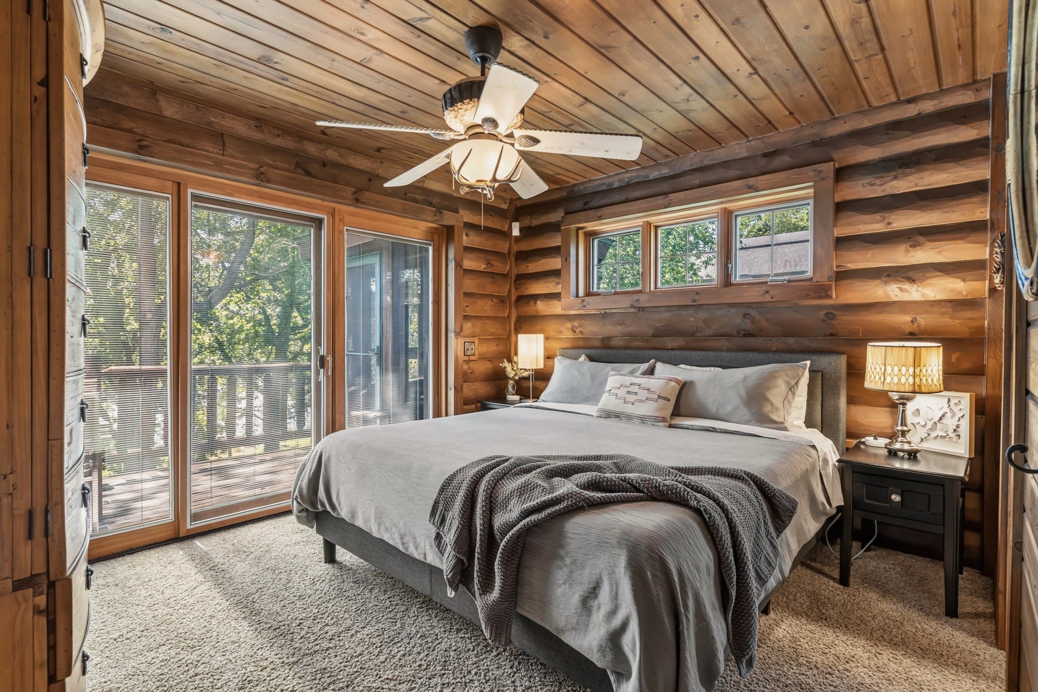 A bedroom with log walls and a plank ceiling: a king bed in grey linen with a knit throw, black nightstands with lamps, a ceiling fan, a row of small windows above the bed, and sliding glass doors onto the deck.