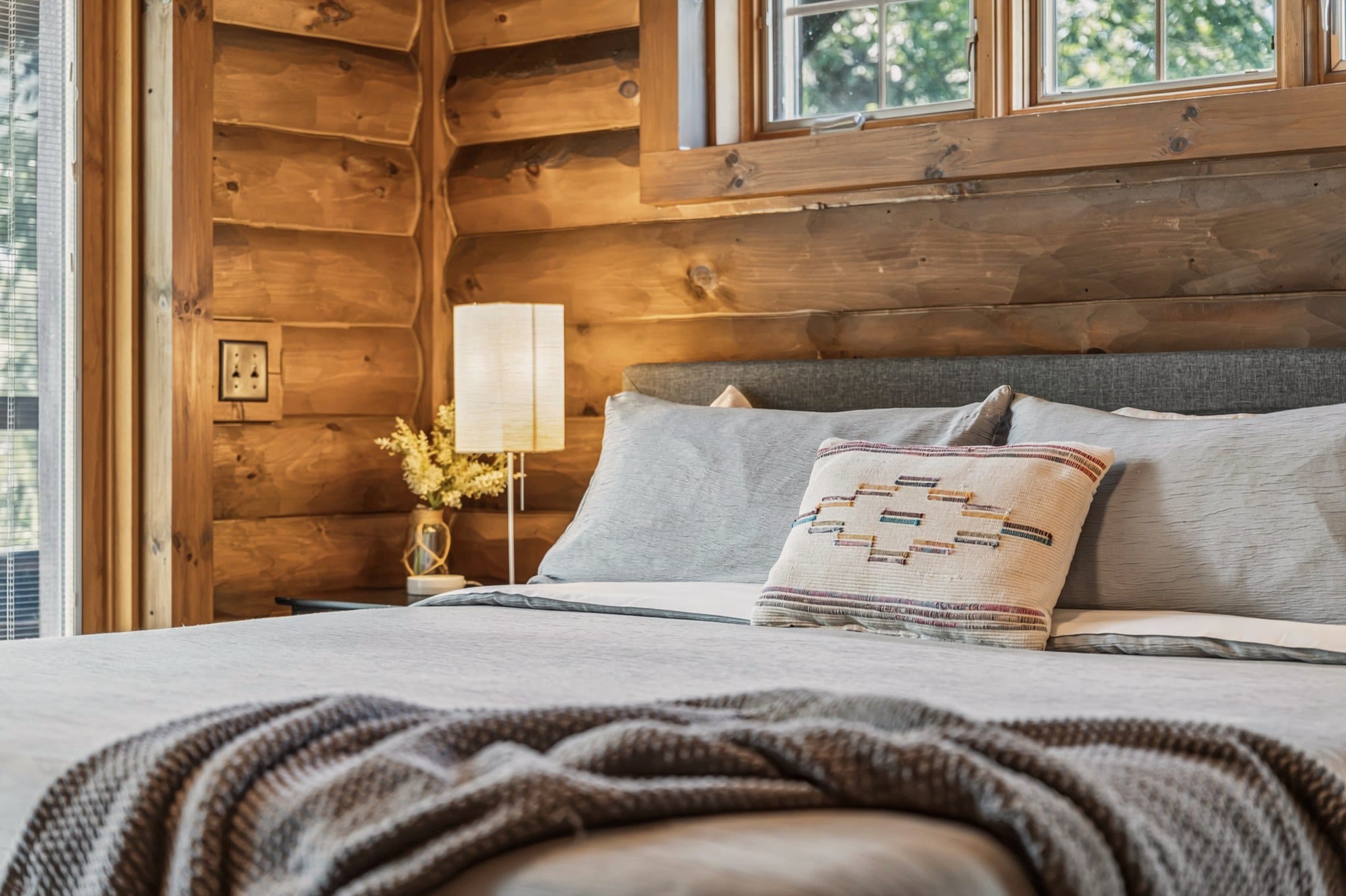 Close view of the same bed: grey linen, a woven throw and a cream cushion stitched with a diamond pattern, against the log wall with a paper-shade lamp and a small vase of yellow flowers.