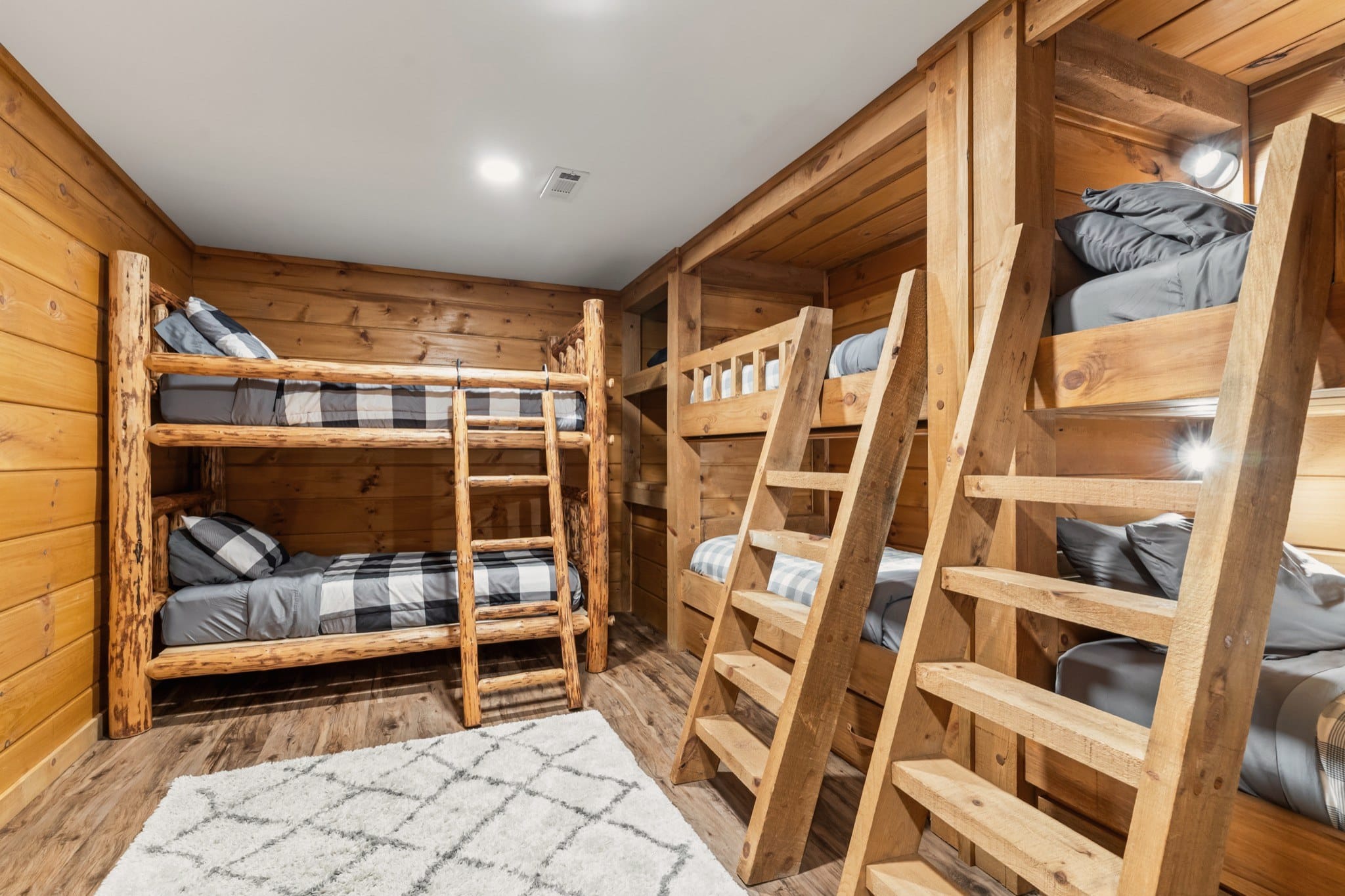 The bunk room: a log bunk bed with a ladder on the left and two built-in timber bunks with ladders and reading lights on the right, all made up in grey and check, over a white shag rug.