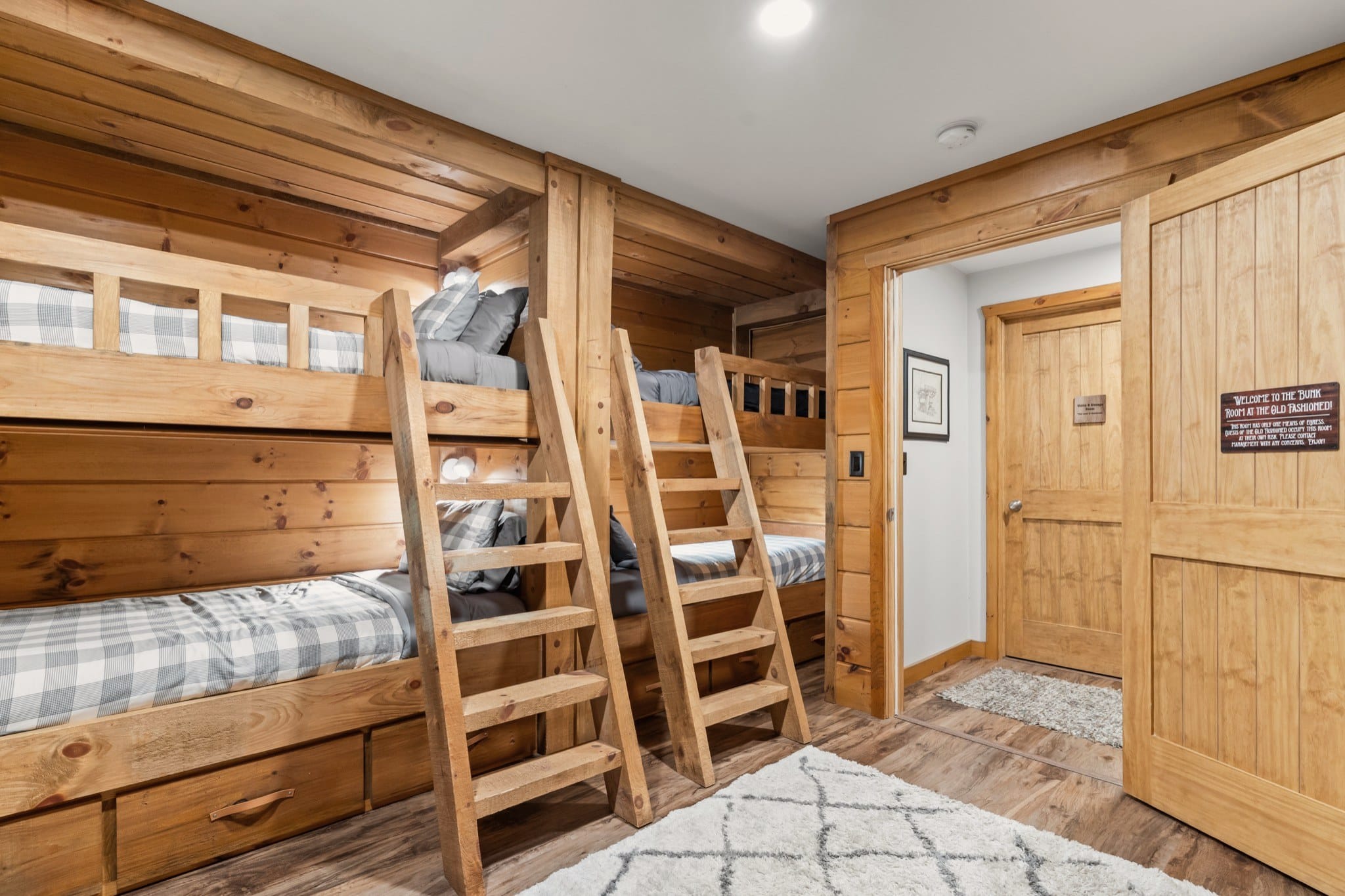 The two built-in bunks with plaid bedding, ladders, reading lights and storage drawers under the lower berths, beside a sliding wooden door with a sign welcoming guests to the Bunk Room at the Old Fashioned.