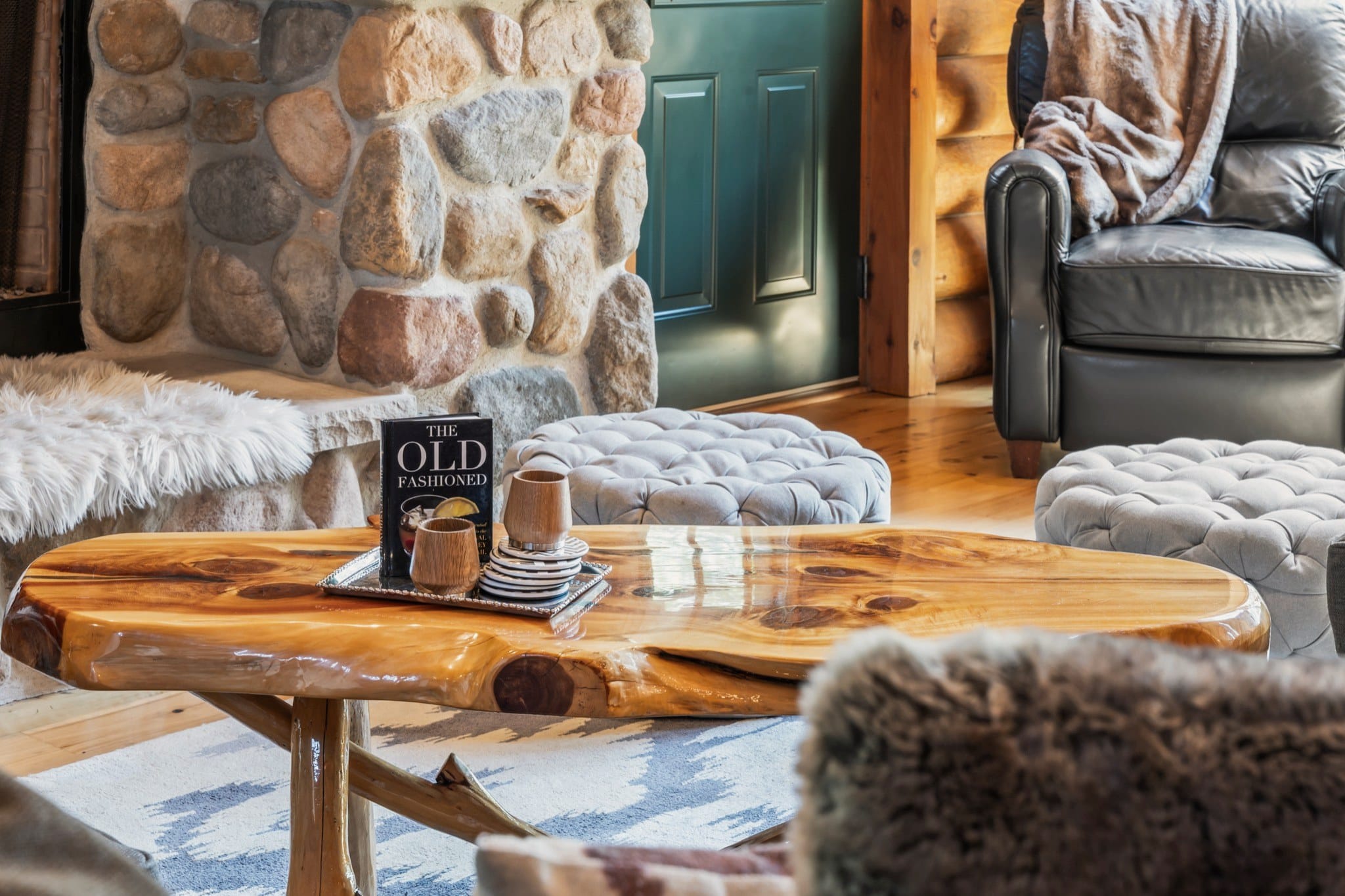 Close view of the polished live-edge coffee table, with a book titled The Old Fashioned and two wooden cups on a tray, a white fur throw on the stone hearth behind it, tufted ottomans and a leather armchair.