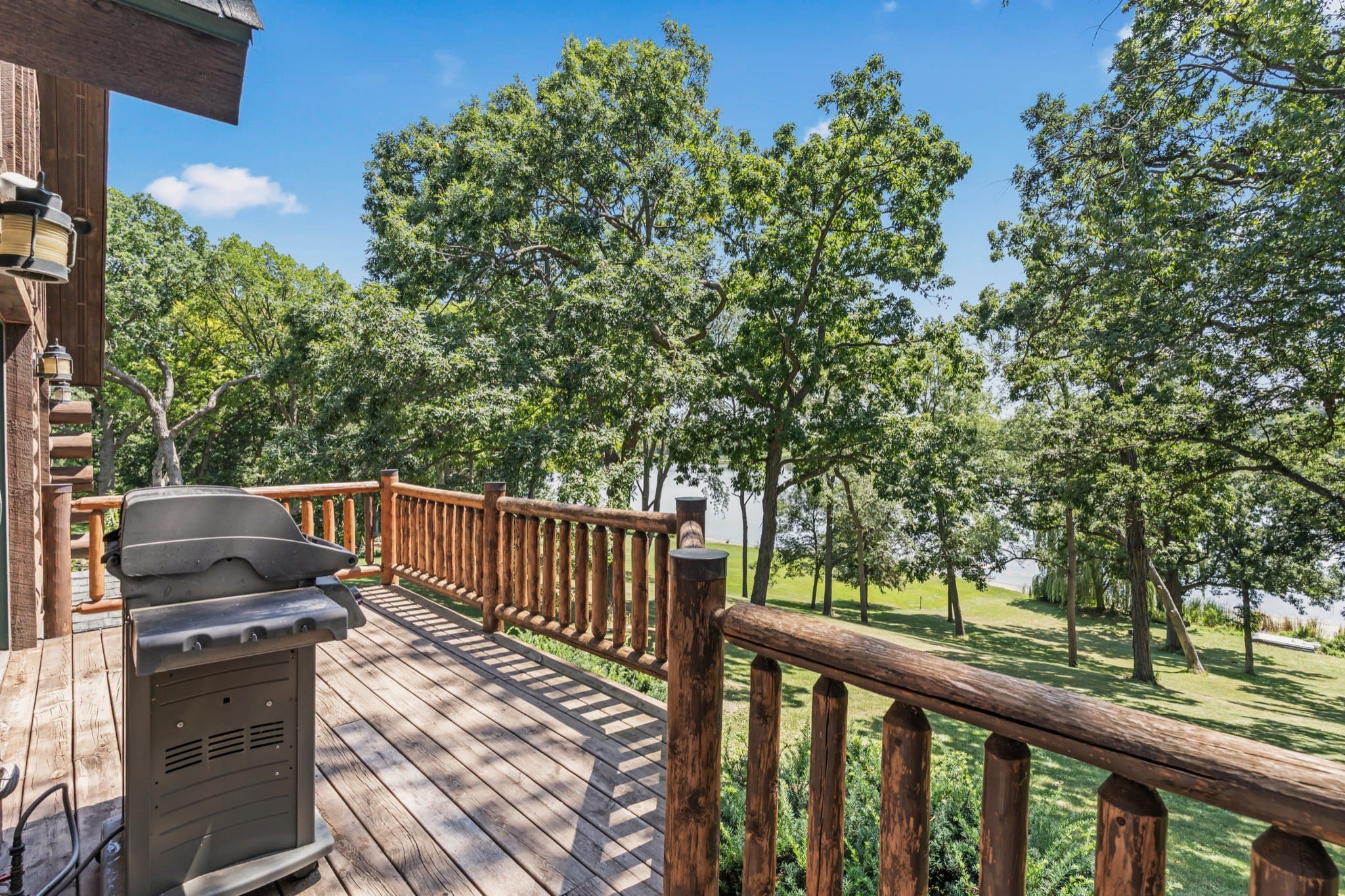 The deck with its log railing and a grill, looking down a lawn shaded by tall trees to Big Twin Lake between the trunks.