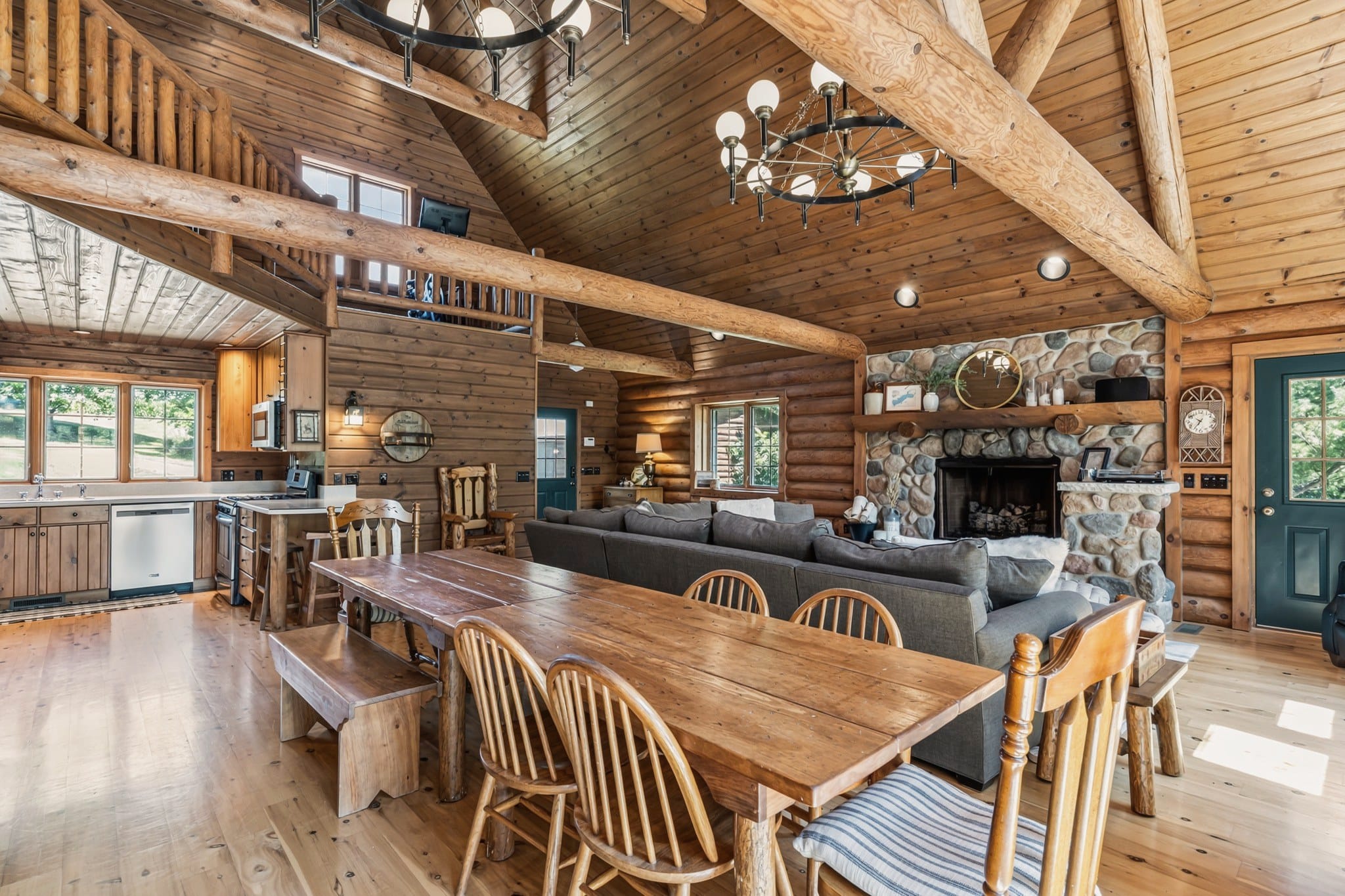 The dining end of the great room: a long plank table with spindle-back chairs and a bench, the grey sectional facing the stone fireplace, the kitchen under the loft on the left, and a ring chandelier with globe lights overhead.