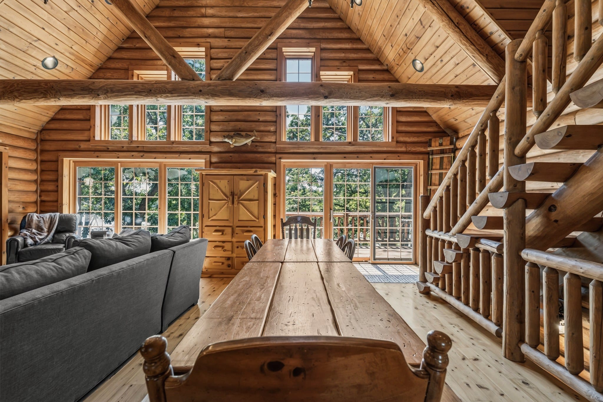 Along the long plank dining table toward the gable wall: tall windows, a sliding glass door to the deck, a wooden armoire, a fish mounted on the wall above the armoire, the grey sectional on the left and the log staircase on the right.