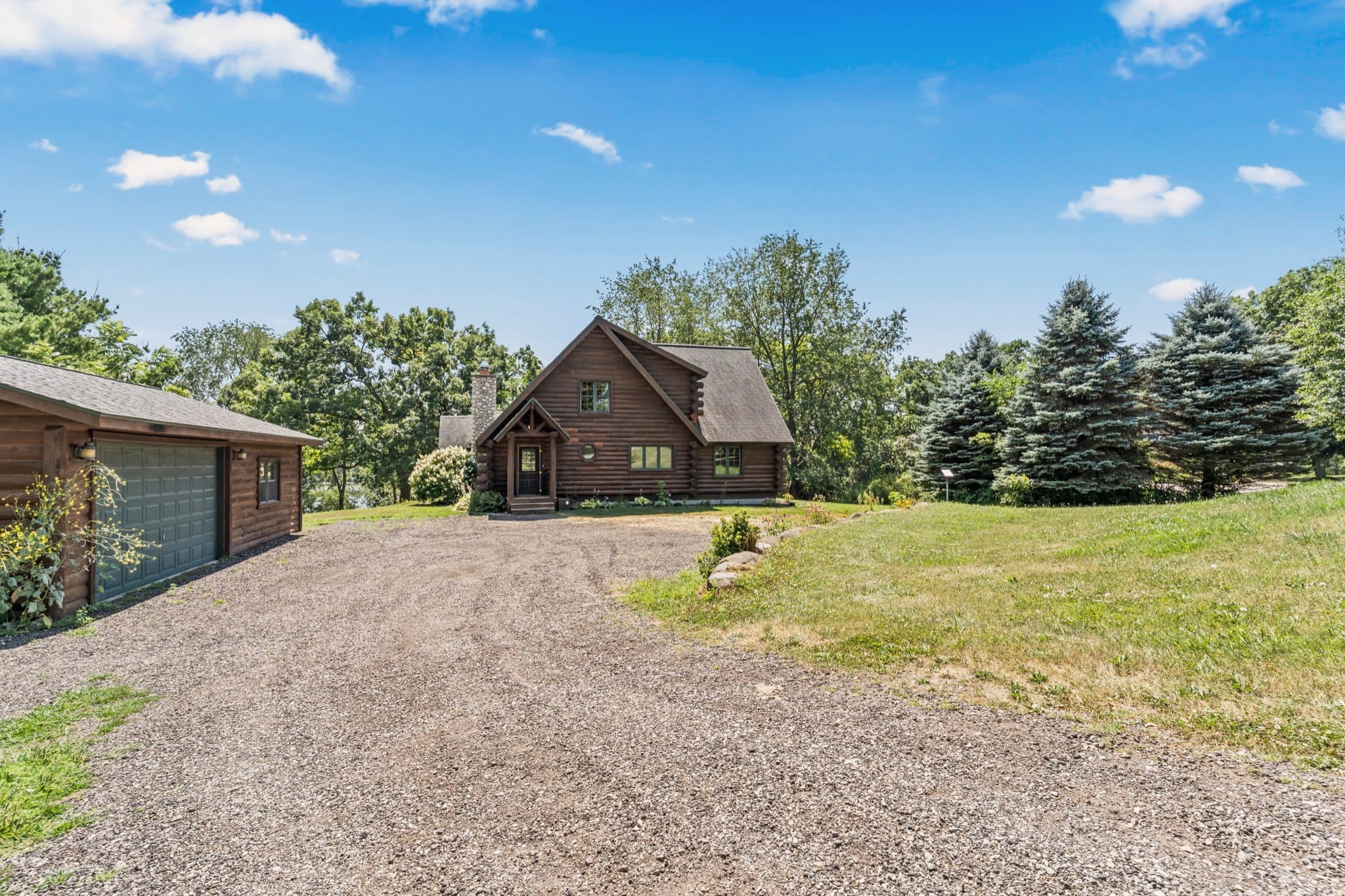 The gravel drive leading to the front of the cabin, past the detached log garage with a green door, with lawn and blue spruces on the right and a glimpse of Big Twin Lake through the trees beside the cabin.
