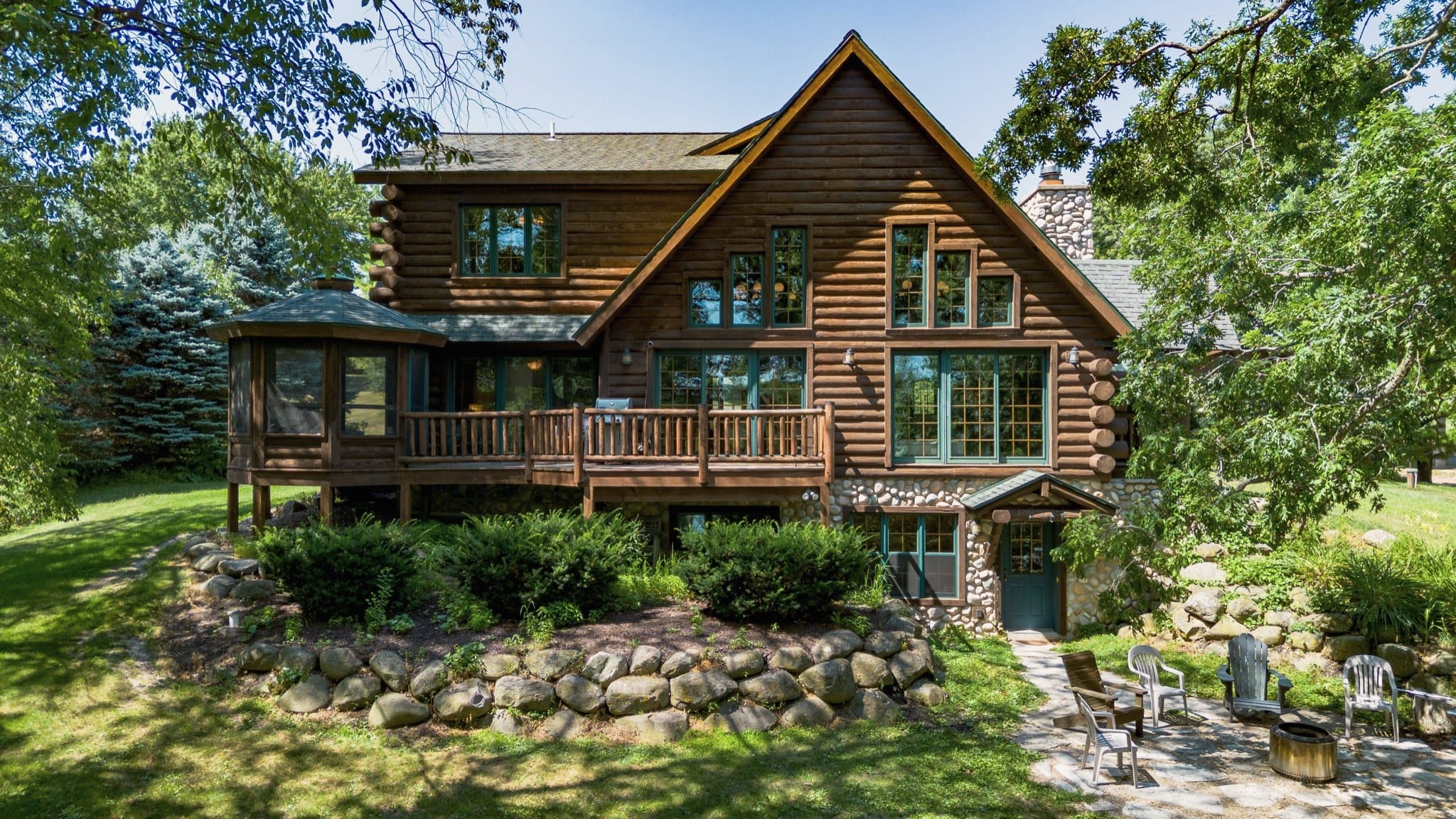 Drone view, low over the lawn, of the back of the cabin: log walls and a gable of tall windows, a long deck with a log railing, a grill and a screened gazebo at one end, a fieldstone lower level with a green walkout door, and a stone patio with Adirondack chairs around a fire pit.