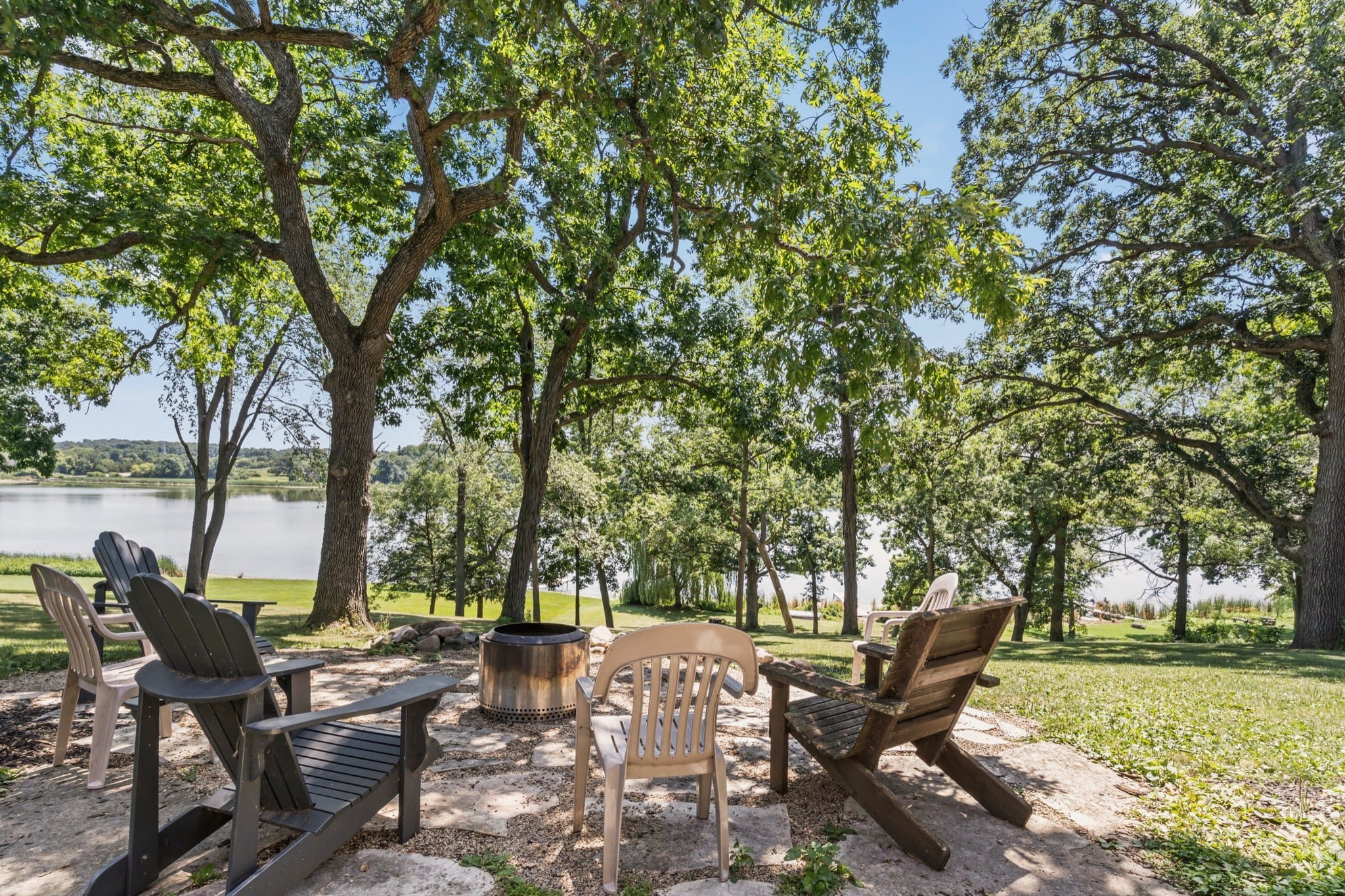 The fire pit on a flagstone patio under tall trees, ringed by Adirondack and patio chairs, with the lawn running down to Big Twin Lake.