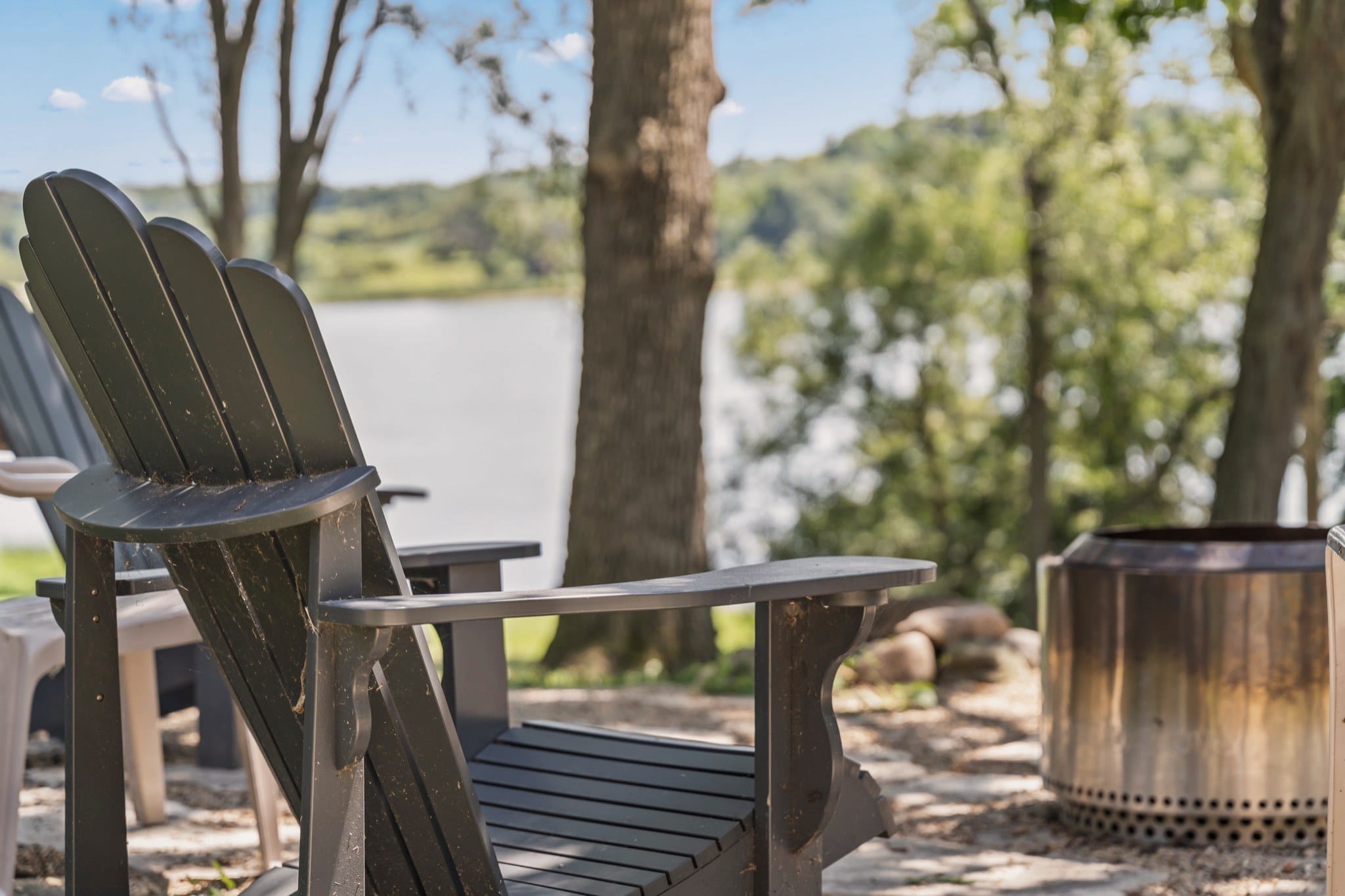 Close view of a grey Adirondack chair beside the steel fire pit, with Big Twin Lake and its wooded far shore out of focus beyond the trees.