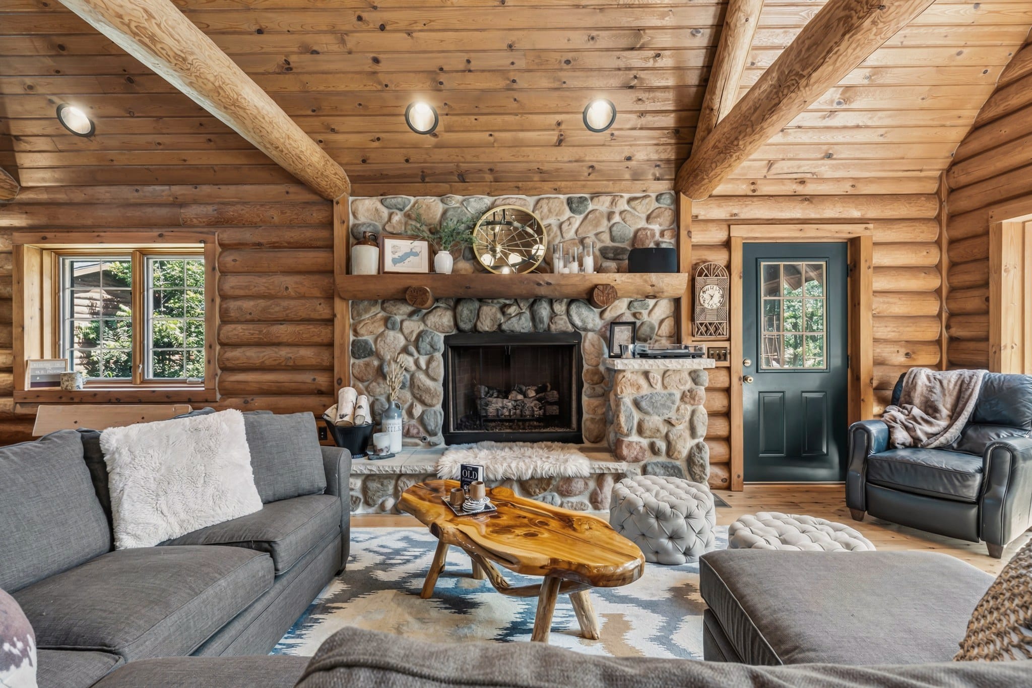 The fieldstone fireplace with a log mantel, seen across a grey sectional and a polished live-edge coffee table, with tufted ottomans, a record player on the stone ledge, a leather armchair and a green door to the outside.