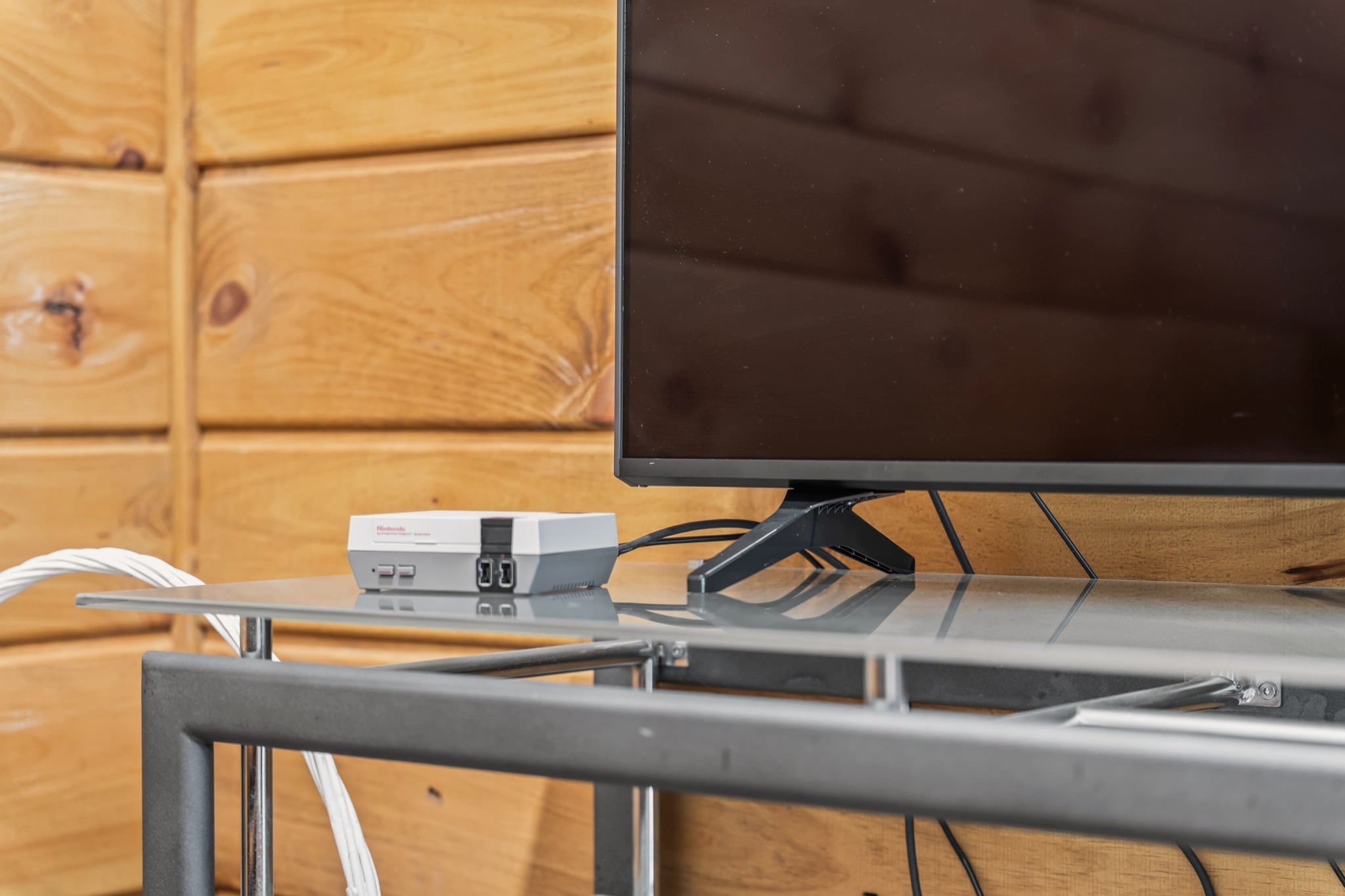 Close view of a small retro game console on a glass and steel TV stand, beside the foot of a flat-screen TV, against a pine plank wall.