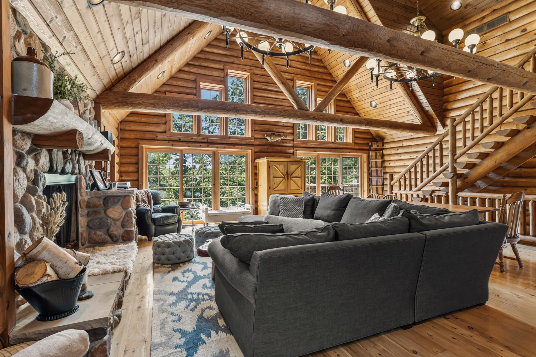 The two-story great room from beside the stone fireplace: a grey sectional, tufted ottomans and a leather armchair under log beams and globe chandeliers, a wall of windows and glass doors to the deck, a wooden armoire, and the log staircase to the loft.