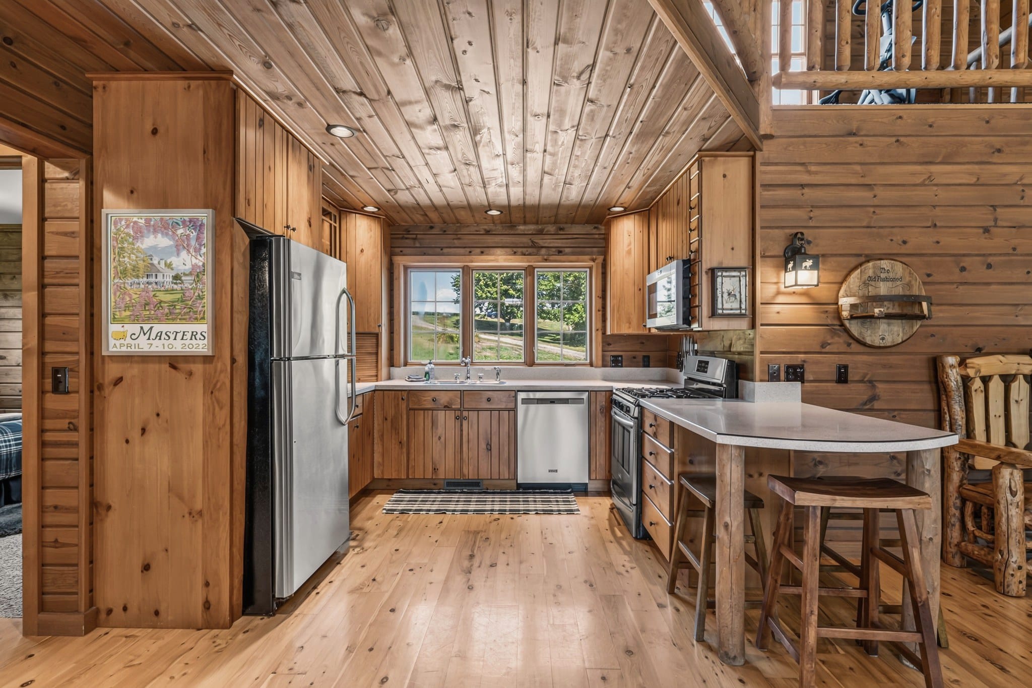 The kitchen: knotty pine cabinets under a plank ceiling, a stainless fridge, range, dishwasher and microwave, the sink under three windows, and a breakfast bar with two wooden stools, with a framed Masters poster and a round wooden sign reading The Old Fashioned on the walls.