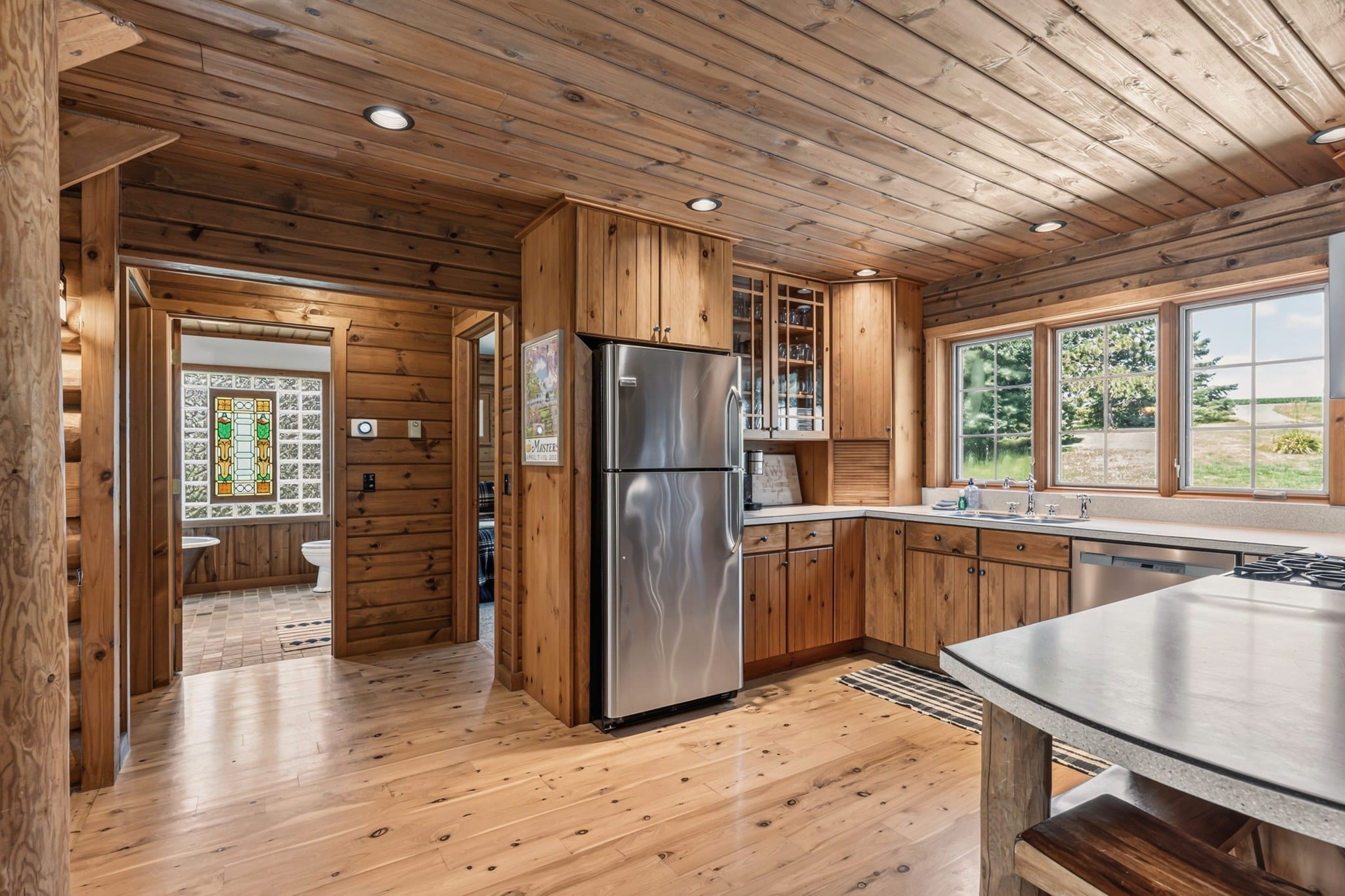 The kitchen from the breakfast bar: a stainless fridge beside a glass-front cabinet of glassware, the counter and sink under three windows, and a hall on the left leading to a bathroom with a stained glass window set in glass block.