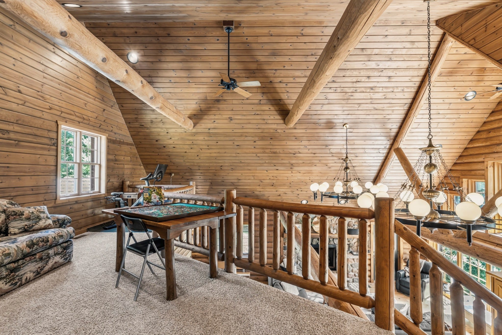 The carpeted loft under the roof's log beams: a patterned sofa, a table with a jigsaw puzzle, an exercise bike by the window and a ceiling fan, with a log railing looking down past the globe chandeliers into the great room.