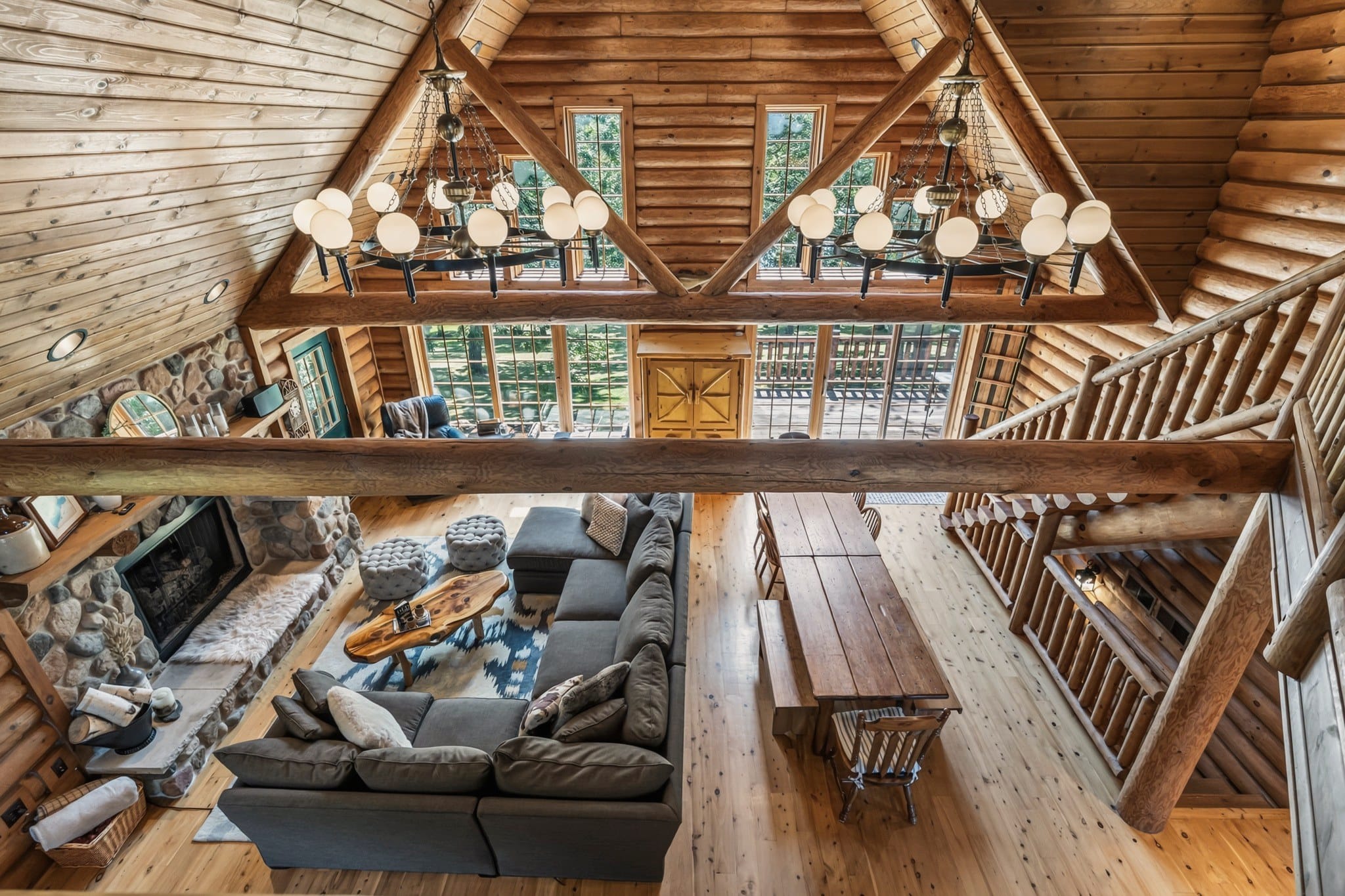 The great room from the loft: the grey sectional and live-edge coffee table by the stone fireplace, the long plank dining table and bench beside the log staircase, and two ring chandeliers hanging in front of the gable windows.