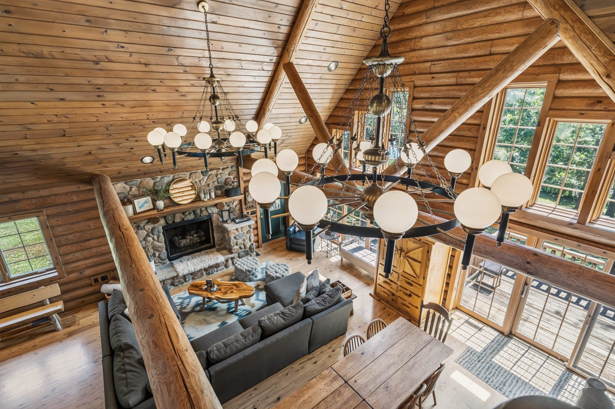 Looking down from the loft past a ring chandelier with globe lights: the stone fireplace with a white fur throw on its hearth, the grey sectional around the live-edge coffee table, the dining table, and glass doors onto the deck.
