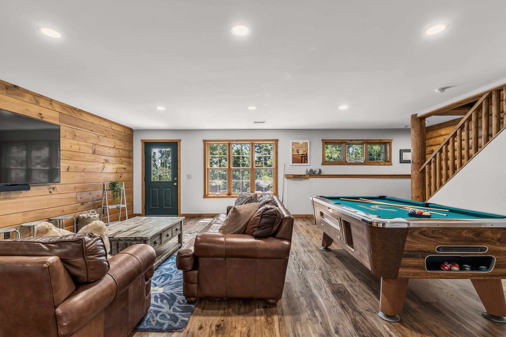 The lower level at the foot of a log staircase: two brown leather sofas and a rustic coffee table facing a large wall-mounted TV, a pool table, and a green door with a window to the outside.