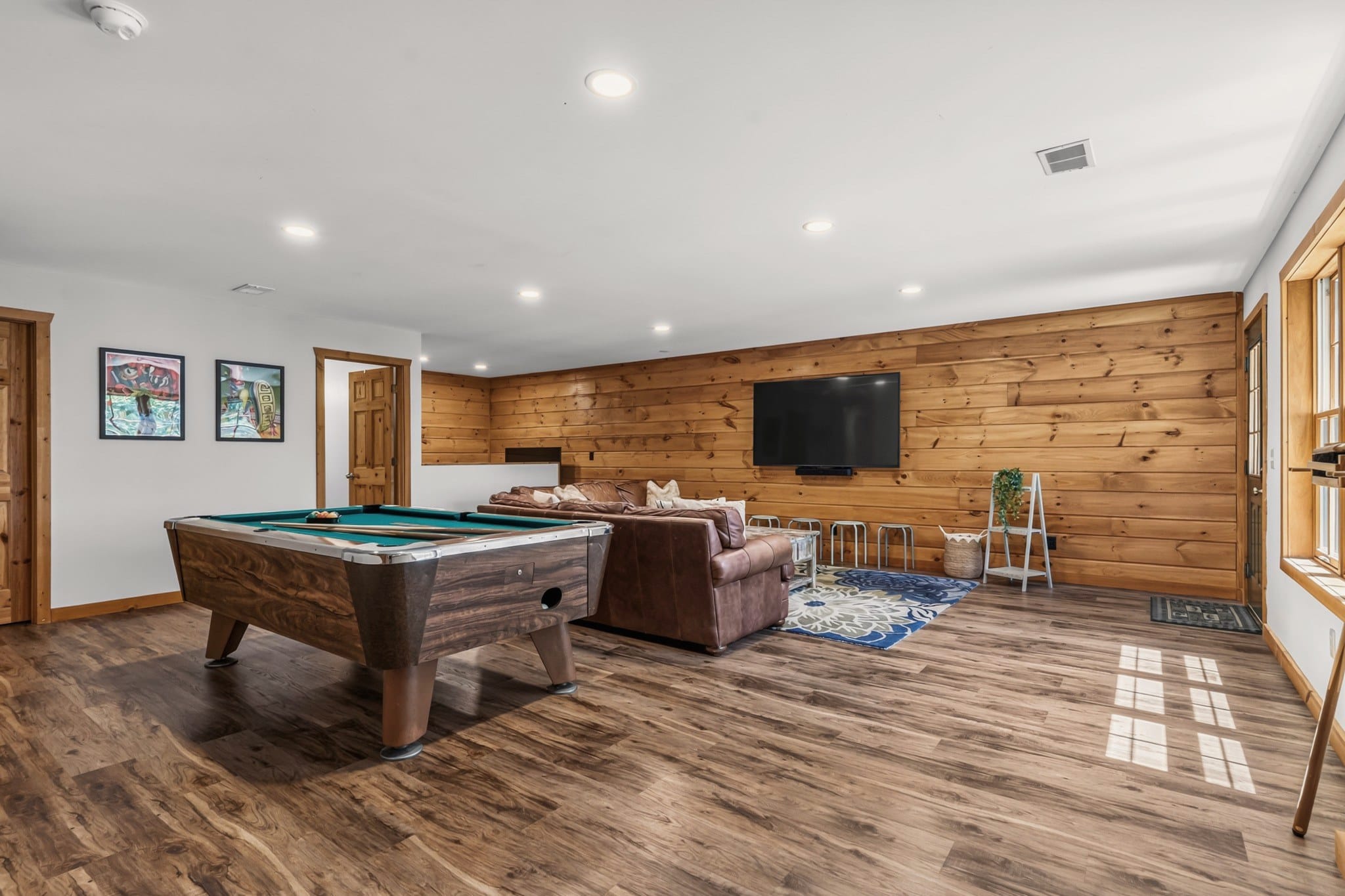 The lower level from the other end: the pool table, a brown leather sectional facing a large wall-mounted TV on a pine plank wall, a row of low stools, framed art prints and wood-look floors.