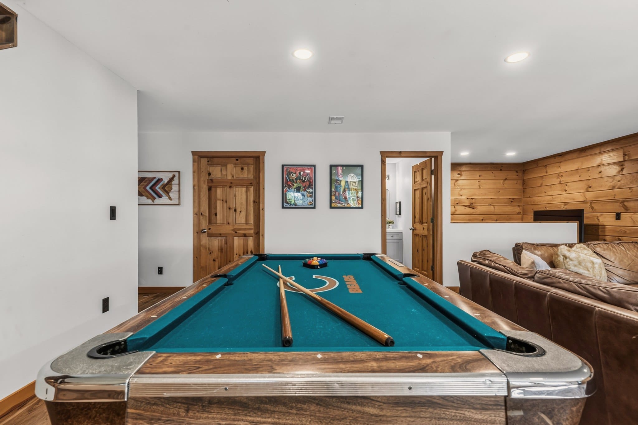 The pool table close up, its teal felt printed with the Chicago Bears logo, two cues and the racked balls, with a brown leather sectional alongside and a door open to a bathroom with a grey vanity.